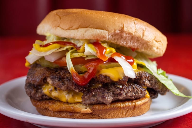 A double cheeseburger with lettuce, tomato, onion, mustard, and ketchup on a white plate.