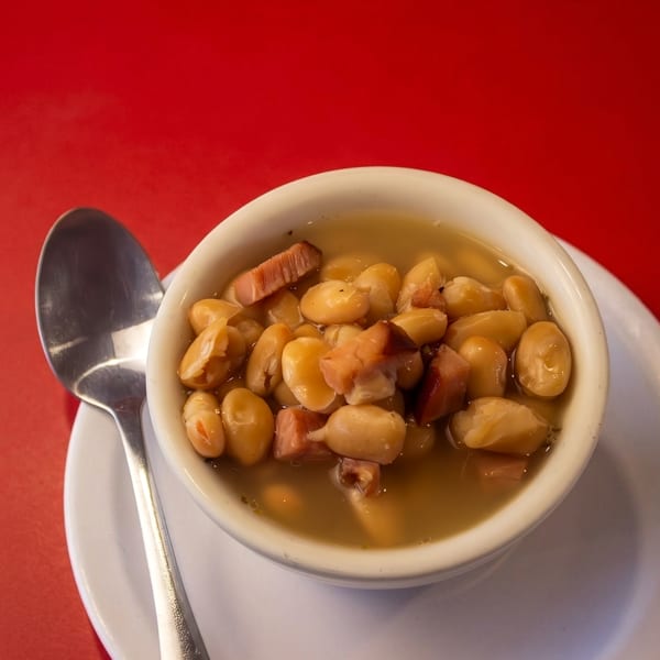 A white bowl of beans and ham in broth, with a spoon, on a red table.
