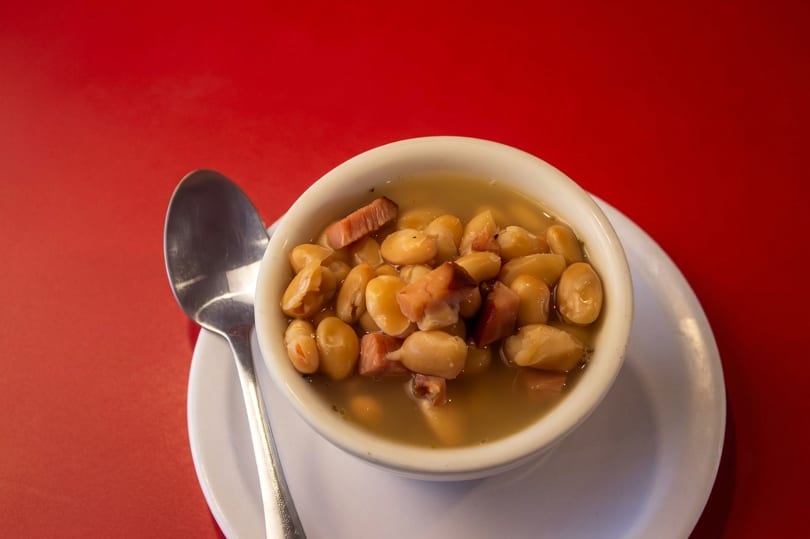 A white bowl of beans and ham in broth, with a spoon, on a red table.
