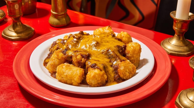 A plate of chili cheese tater tots on a red table with brass candle holders.