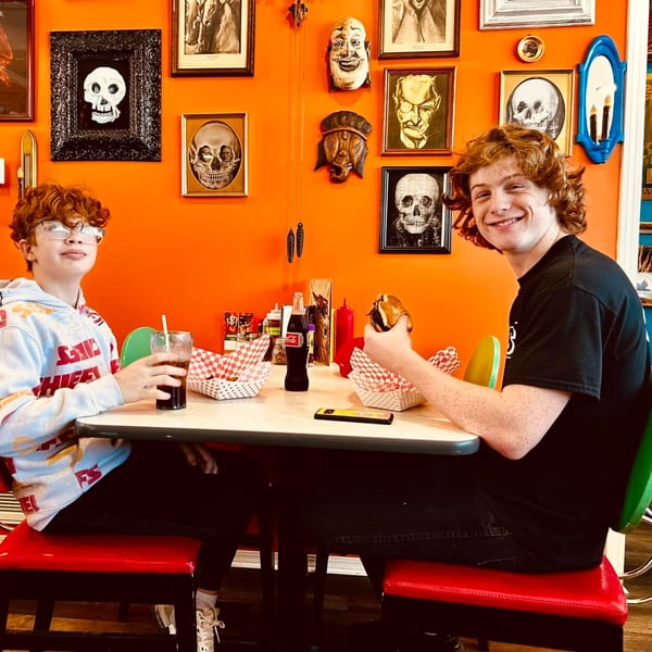 Two boys sit at a diner table with burgers and drinks, against an orange wall with skull art.
