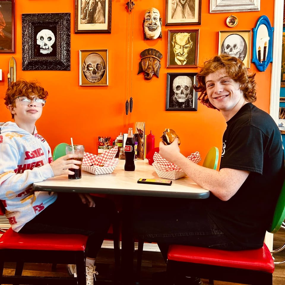 Two boys sit at a diner table with burgers and drinks, against an orange wall with skull art.