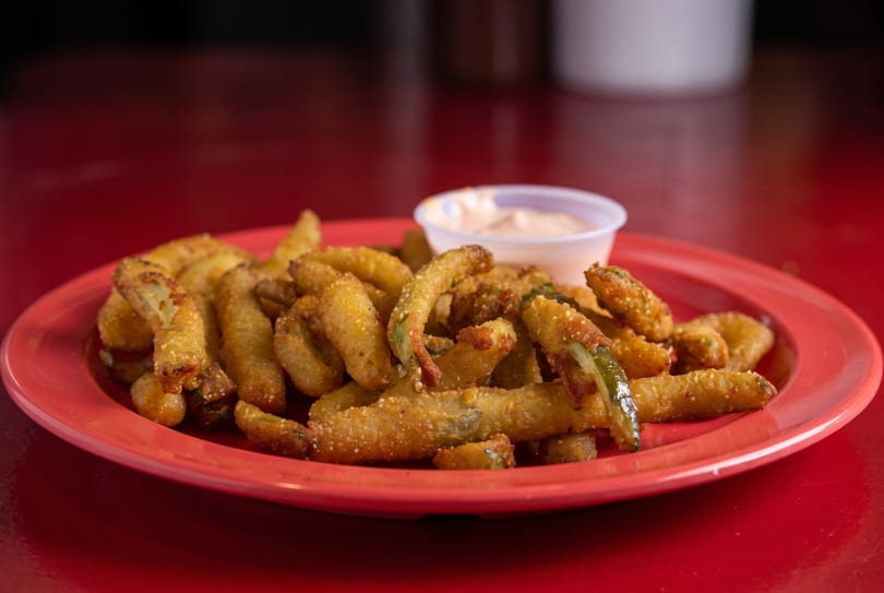 Crispy fried pickle spears on a red plate with a side of dipping sauce.